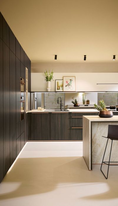 Moderne Küche mit dunklen Holzschränken, einer zentralen Insel und Pflanzen auf der Arbeitsplatte. Helle Wände und Decke schaffen eine einladende Atmosphäre.