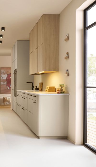 Moderne Küche mit hellen Möbeln, Holzregalen und einem großen Fenster. Ein Teppich liegt auf dem Boden, und es gibt Pflanzen im Raum.